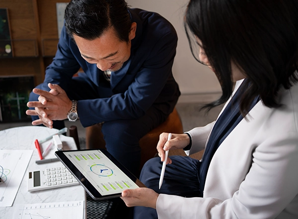 A candid, high-angle shot shows two business professionals collaborating over a tablet. A person with dark hair, wearing a white blazer, is seated in the foreground on the right, holding the tablet and a white stylus, pointing at the screen which displays a graph and a clock icon. Another person, a man with dark hair and a dark blue suit, is leaning over from the left, looking down at the tablet with a slight smile or thoughtful expression, his hands clasped in front of him. On the table, a white calculator and a red pen are visible. The background is slightly blurred, showing what appears to be an indoor office or lounge setting with books on a shelf.
