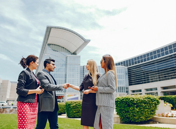 Four well-dressed individuals are standing outdoors, seemingly engaged in a business interaction, with modern buildings and a clear sky in the background. On the left, a woman in a black blazer and a red and white polka dot skirt stands next to a man in a gray suit, who is shaking hands with a woman in a dark dress. This woman has long blonde hair. To her right, another woman with long brown hair and sunglasses is wearing a gray plaid suit. Lush green grass and manicured bushes are in the foreground. The architecture behind them suggests a corporate or government setting.