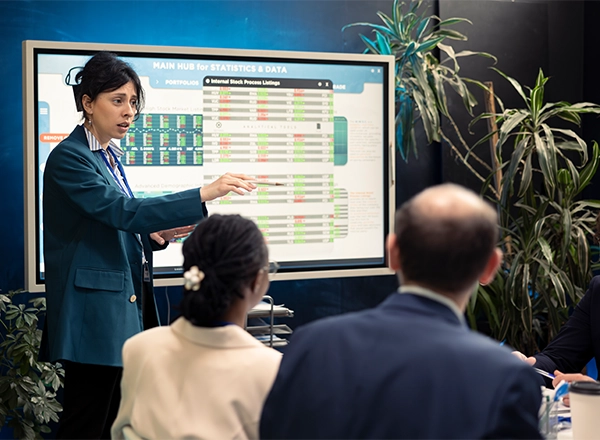 A woman with dark hair, wearing a teal blazer over a striped shirt, is presenting in front of a large screen displaying "MAIN HUB FOR STATISTICS & DATA" and complex charts/tables. She is pointing at the screen with a pen and speaking. In the foreground, the backs of two people's heads are visible, indicating an audience. Lush green plants are visible on either side of the screen.