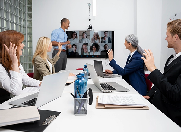 A diverse group of professionals are in a meeting room, some waving at a large screen displaying a video conference call, with a presenter standing by the monitor.