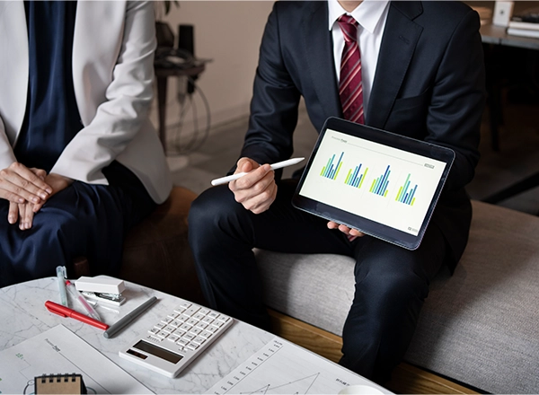 A person in a suit and tie holds a tablet displaying bar graphs, while another person sits beside them and a calculator and pens are on the table.