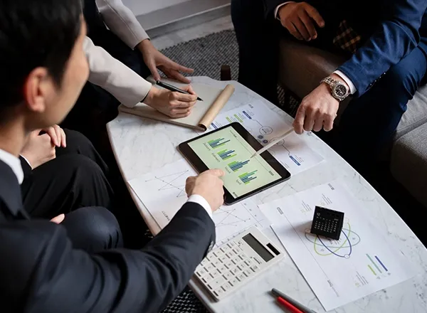 A high-angle shot captures three people gathered around a marble-top coffee table, reviewing business documents and a tablet displaying bar graphs. One person points to the tablet with a stylus, another takes notes in a notebook, and a third observes. A calculator, pens, and a small black clock are also on the table.