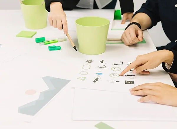 A high-angle shot captures three people gathered around a white table, focusing on design elements. Two hands are visible in the foreground; one points with a finger at a sheet of paper displaying various icons related to recycling and energy, while the other holds a pencil, pointing towards a light green pot. Another person's hand is visible in the background. Green office supplies like markers and sticky notes are scattered on the table.