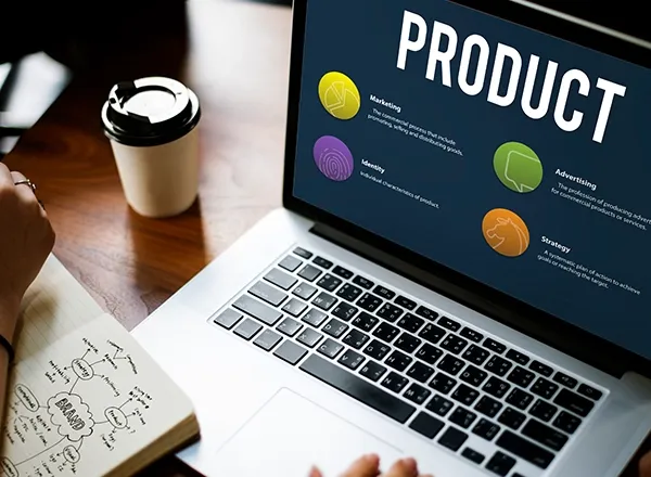 A person is seated at a wooden desk, looking at a laptop displaying a presentation slide with the large word "PRODUCT" at the top. Below "PRODUCT" are four sections: "Marketing," "Identity," "Advertising," and "Strategy," each with an icon and a brief description. To the left of the laptop, there's an open notebook with a "BRAND" mind map, and a white disposable coffee cup with a black lid. The person's hands are visible near the laptop's keyboard.