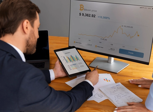 A man in a suit sits at a wooden desk, holding a tablet displaying bar graphs while looking at a large monitor showing a "Bitcoin price" chart with an upward trend. The monitor shows "Bitcoin price $9,362.92 +10,569%". On the desk, there are papers with charts, a notebook with handwritten notes, and a laptop. A woman's hand is visible on the right side of the image, near the notebook.