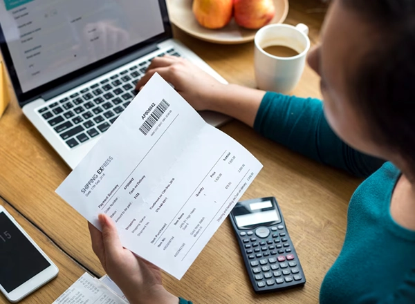 A high-angle shot captures a person from over their shoulder as they work on a wooden desk. The person is holding a document labeled "SHIPPING EXPRESS" with a barcode and line items, while their other hand is typing on a silver laptop. To the right of the laptop, a white mug with coffee and a plate with two oranges are visible. Below the document, a black calculator is on the desk, and a white smartphone is to the left. A receipt is partially visible near the smartphone.