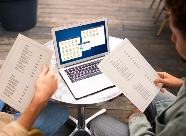 A high-angle shot shows two people sitting at a small, round table, both holding up white papers with lists of text. Between them, a silver laptop is open on the table, displaying a screen with multiple windows that appear to show a calendar or scheduling interface. The person on the left is wearing light-colored pants, while the person on the right is wearing dark pants and a light-colored top. The floor is made of wooden planks, and blurred elements of an indoor setting are in the background.