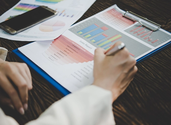 A person's hands are visible in the foreground, holding a silver pen and writing or marking on a document labeled "Sales Report." This document is attached to a blue clipboard and displays various charts and graphs, including bar charts and line graphs, with red and blue elements. Another document with pie charts and a smartphone are visible in the top left background on a dark wooden surface. The lighting is somewhat dim, with a focus on the hands and documents.