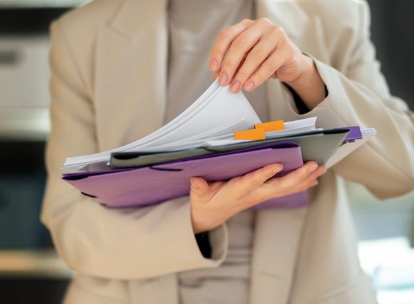 A person wearing a light-colored blazer is holding a stack of documents and folders. Their hands are in the process of organizing or retrieving papers from the folders. There's a purple folder, a dark gray or green folder, and white papers visible, some of which have orange paper clips or tabs. The background is blurred, suggesting an indoor office or professional setting.