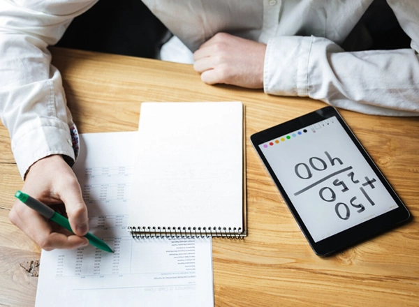 A person wearing a white long-sleeved shirt is seated at a wooden table, engaged in work that involves a document, a notebook, and a tablet. In the foreground on the left, one hand holds a green pen, marking or writing on a paper with lines of text and numbers. Next to it, an open spiral-bound notebook with blank pages is visible. To the right, a black tablet displays handwritten numbers and symbols, including "