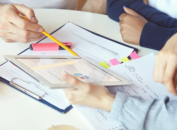A top-down shot shows two people at a light-colored table, engaged in a discussion over documents and a tablet. On the left, a person's hands are visible, holding a yellow pencil and pointing towards the tablet or papers. On the right, another person's hands are holding a tablet, which displays colorful charts. Various papers and clipboards are scattered across the table, some with neon pink, yellow, and green sticky notes attached. A pink highlighter also lies on the table.