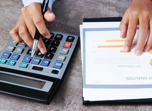 A high-angle shot captures two people, one with light skin and the other with dark skin, collaborating over documents and a laptop on a light wooden table. The person with light skin, wearing a light blue long-sleeved shirt, is pointing with a pen towards the laptop screen, which displays a bar graph. The person with dark skin, wearing a white long-sleeved shirt, is also holding a pen and pointing at a physical document on the table that features colorful charts and graphs. Various other papers with charts and a calculator are scattered on the table, indicating a business or financial analysis discussion.