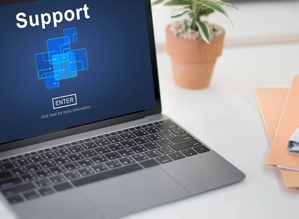 A close-up shot shows a laptop computer displaying the word "Support" in white text at the top left of its screen. Below "Support" is a graphic of interconnected blue squares, and in the center of the screen, a white "ENTER" button is visible within a black rectangle, with "click here for more information" written beneath it. The laptop is open on a white surface, with part of a potted plant and some folders visible in the blurred background to the right.