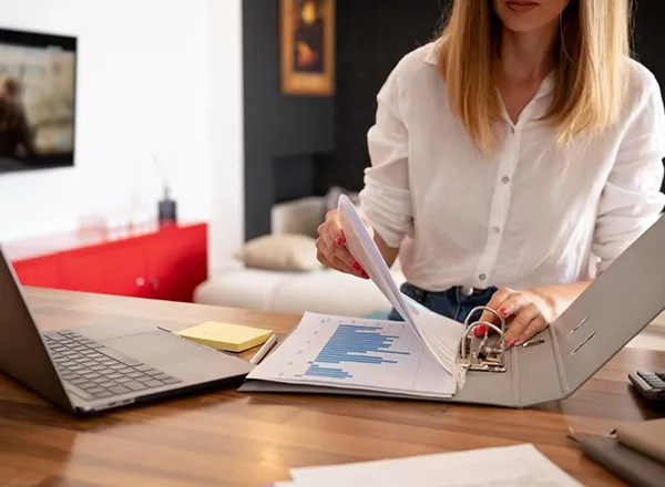 A person with light-colored hair, wearing a white long-sleeved shirt and blue jeans, is standing at a wooden table, flipping through papers inside a gray three-ring binder. The papers include a sheet with a blue bar graph. To the left of the binder, a silver laptop is open on the table, along with a yellow sticky note. In the background, a TV is mounted on a white wall, and a red cabinet and a light-colored couch are partially visible, suggesting a home or office setting. The focus is on the person's hands and the documents.