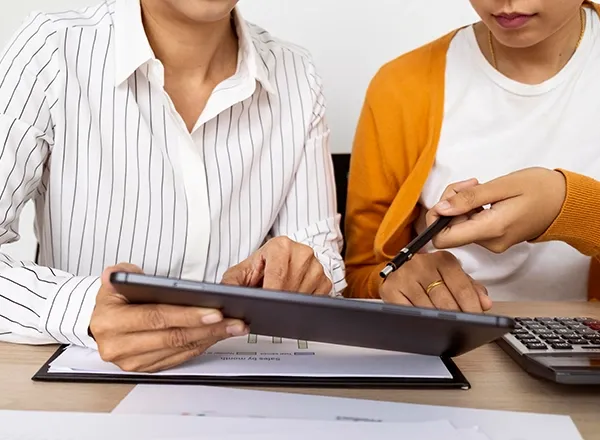 A close-up shot shows two individuals, likely women, collaborating on a task involving a digital tablet and documents. The person on the left is wearing a white pinstriped shirt and holding a black tablet horizontally, seemingly displaying a graph or chart. The person on the right, wearing a white t-shirt and an orange cardigan, is pointing at something on the tablet or a document with a black pen. A calculator is partially visible on the right, and papers on a clipboard are under the tablet, suggesting a business, financial, or educational discussion.