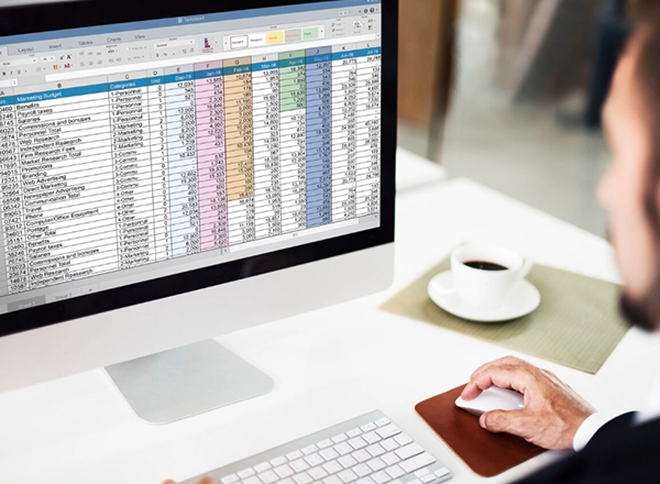 A professional setting is depicted with a person, seen from the shoulder, using a desktop computer. The computer screen displays a detailed spreadsheet, likely for financial data or budgeting, with numerous rows and columns of numbers and categories such as "Marketing Budget," "Personal," and "Research." The person's hands are visible, one on a white mouse with a brown mousepad, and the other on a white keyboard. To the right of the monitor, a white cup of coffee sits on a green placemat. The background is blurred, suggesting an office environment.