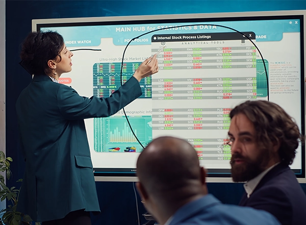 A businesswoman points at a large screen displaying "Internal Stock Process Listings" and other financial data to colleagues.