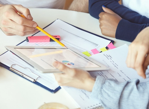 A top-down shot shows two people at a table, looking at documents and a tablet. One person's hands are visible on the left, holding a yellow pencil and pointing to something on a document or the tablet. Another person's hands are visible on the right, resting on the table and holding the tablet. Several papers with writing and graphs are spread across the table, along with a pink highlighter.