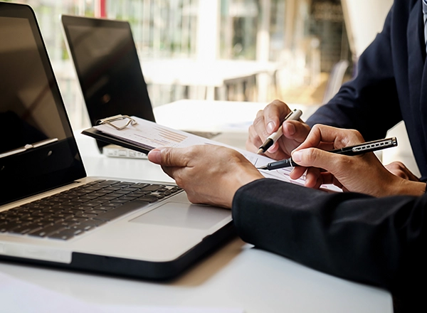 Two people are collaborating at a desk, reviewing documents and using laptops.
