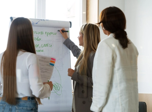 Three women are in an indoor setting, possibly an office or classroom, facing a white flip chart. A woman with long blonde hair, wearing a gray blazer, is writing on the flip chart with a marker. Her back is partially to the viewer, but her profile is visible. To her left, a woman with long dark hair, seen from behind, is holding papers with colorful tabs. To her right, another woman with dark hair, also seen from behind, is wearing a light-colored jacket. The flip chart has some green writing and diagrams on it.